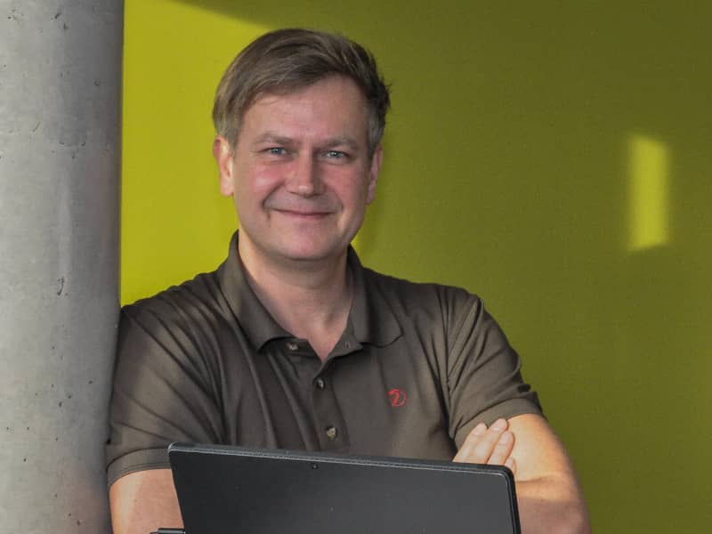 steumawi – Mathias Winkler Das Bild zeigt den Dipl. Kfm. Mathias Winkler, der freundlich und selbstbewusst in die Kamera lächelt. Er lehnt sich leicht an eine graue, raue Säule oder Wand auf der linken Seite an. Er trägt ein dunkelbraunes oder olivgrünes Poloshirt, auf dem ein kleines, rotes Emblem (möglicherweise ein Logo) zu erkennen ist. Seine Arme sind vor der Brust verschränkt, und er hält einen geschlossenen Laptop oder ein Tablet vor sich. Der Hintergrund ist durch eine leuchtend gelb-grüne Wand dominiert, die einen starken Kontrast zum dunklen Hemd bildet.